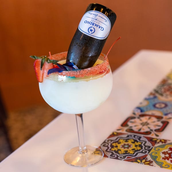 A large margarita with a mini Gambino Cuvee bottle, strawberry, and cherry on a white table.