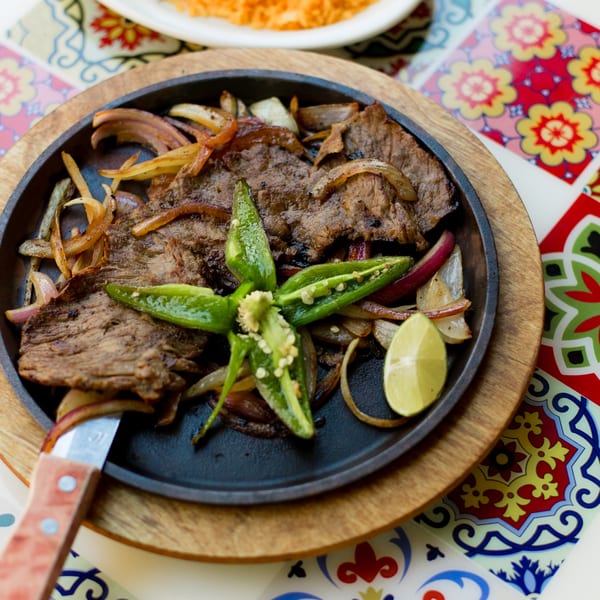 Sizzling El Paso Tacos & Tequila fajitas with steak, onions, jalapeños, and lime on a colorful table.