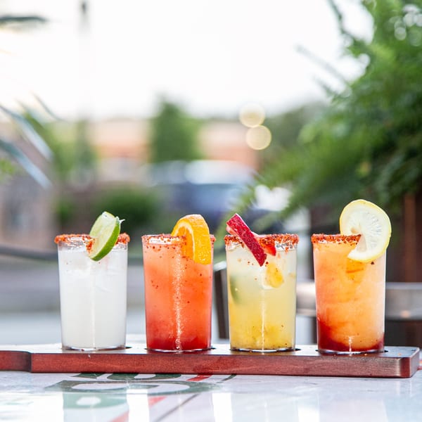 Four colorful El Paso Tacos & Tequila margaritas with fruit garnishes on a wooden flight board.