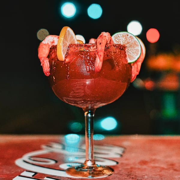 A vibrant red michelada cocktail, garnished with shrimp, lime, and lemon slices, against a dark, blurred background.